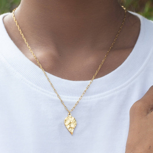 Gold necklace with a leaf-shaped pendant worn by a person in a white shirt.