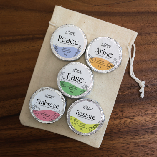 Five small round silver foil-wrapped shower steamers labeled Peace, Arise, Ease, Embrace, and Restore from Simply Healthy, arranged on a cream drawstring pouch over a wooden surface.