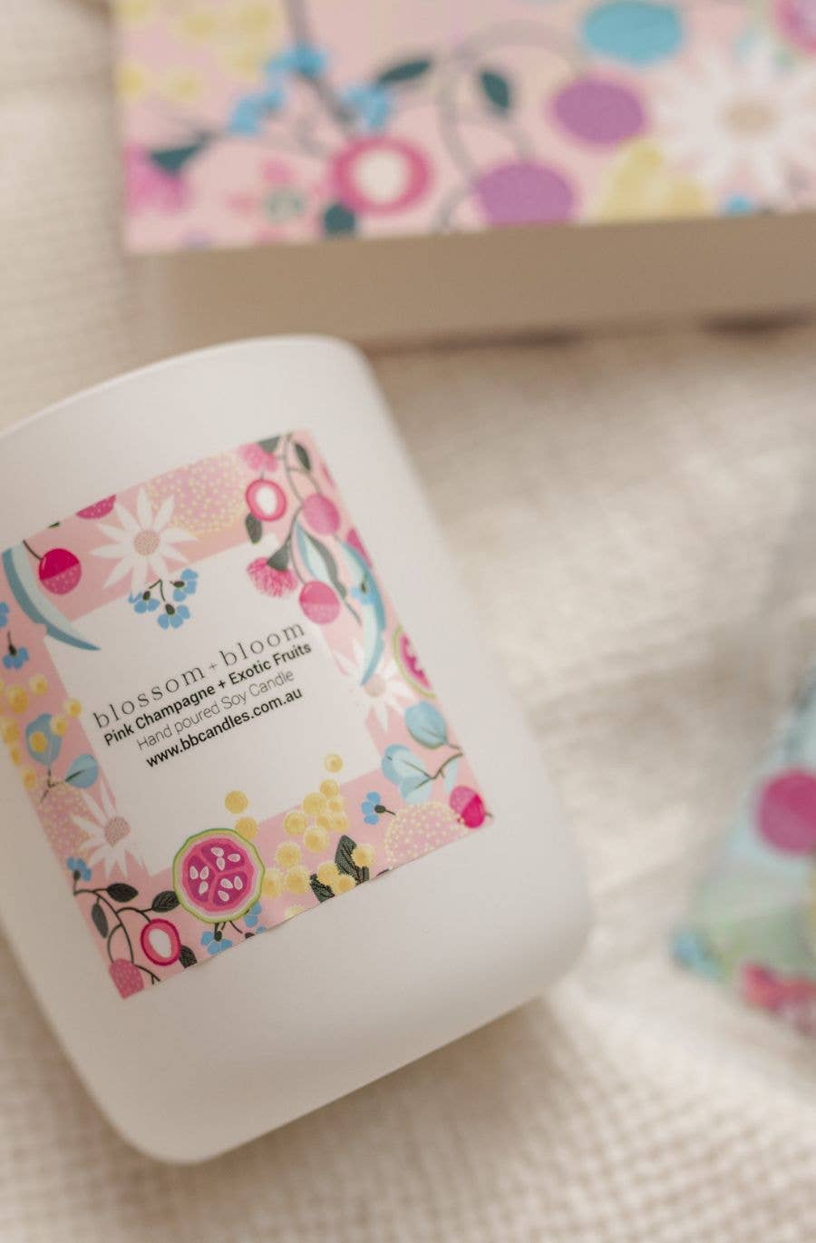 A white candle jar with a colourful floral label featuring the name " Blossom and Bloom," placed on a textured fabric surface. A matching floral-patterned box is partially visible in the background.