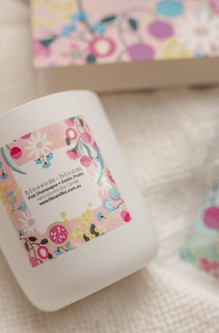 A white candle jar with a colourful floral label featuring the name " Blossom and Bloom," placed on a textured fabric surface. A matching floral-patterned box is partially visible in the background.