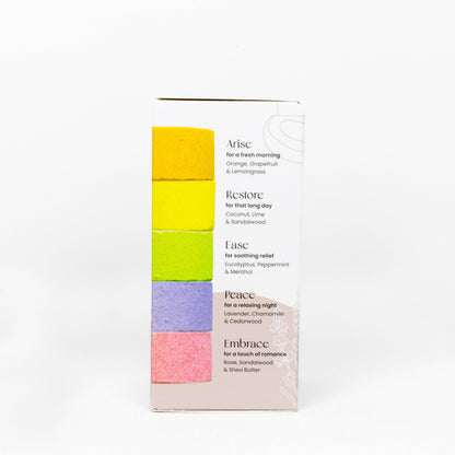Shower Steamer Treat Pack box standing upright on a white background. The front of the box shows five stacked color blocks in yellow, lime green, purple, and pink on a pale blush base, with product names Arise, Restore, Ease, Peace, and Embrace printed beside them.