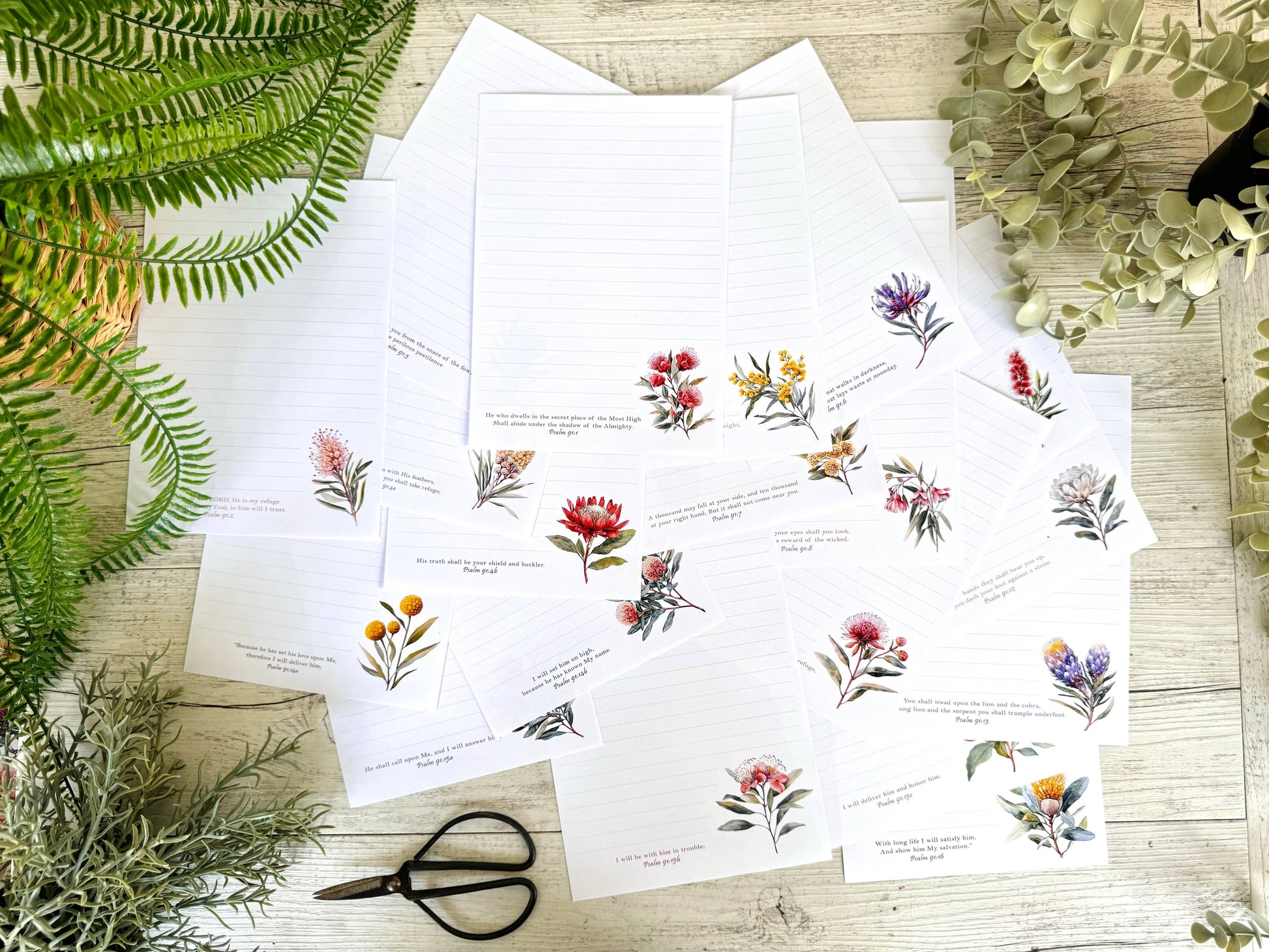 Sheets of paper with scriptures written on them and floral illustrations on a wooden surface with greenery.
