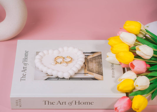Styled arrangement on a pink background featuring a heart shaped bubble trinket dish holding a pair of gold earrings. Yellow and pink tulip flowers are placed on the right side, adding a vibrant touch.
