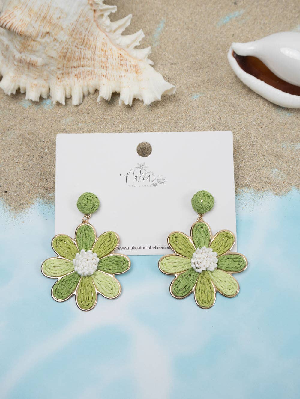 Green raffia daisy flower earrings with white centres on display card, beach background.