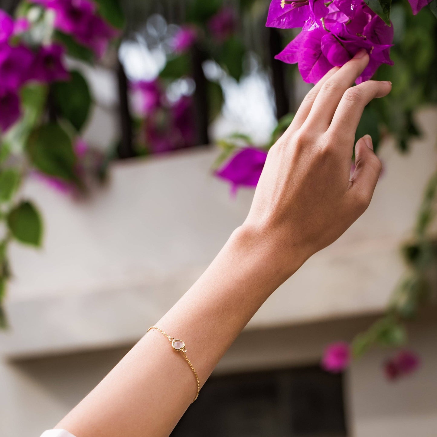 A close-up lifestyle shot of a person's hand and forearm wearing a delicate gold chain bracelet with a small pink glass stone, set against a soft-focus background of purple flowers and green leaves.
