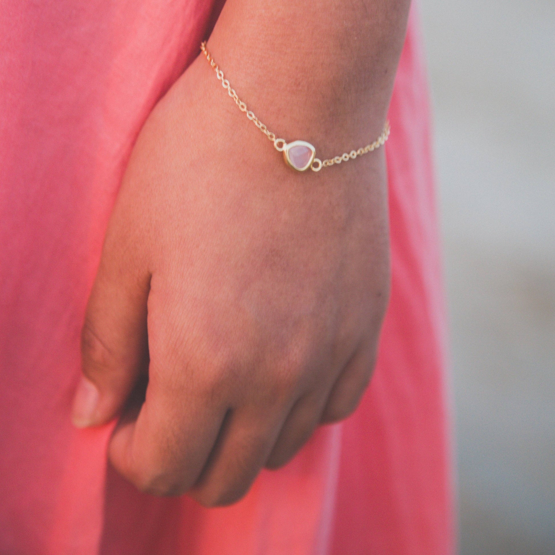 A delicate gold chain bracelet with a small teardrop-shaped pale pink stone charm, worn on a person's wrist. The person is wearing a pink dress, and the background is softly blurred.