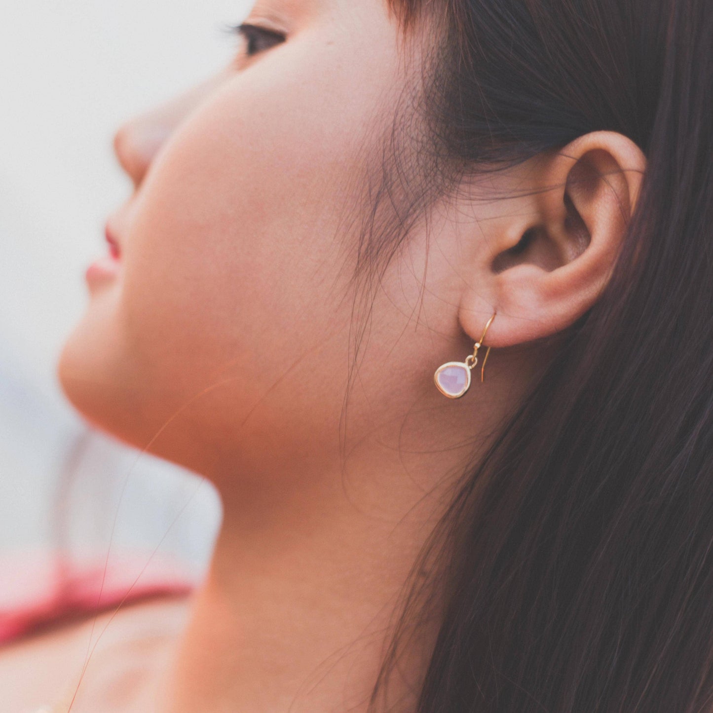 A close-up side profile of a woman wearing a small gold dangle earring with a teardrop-shaped pale pink stone.