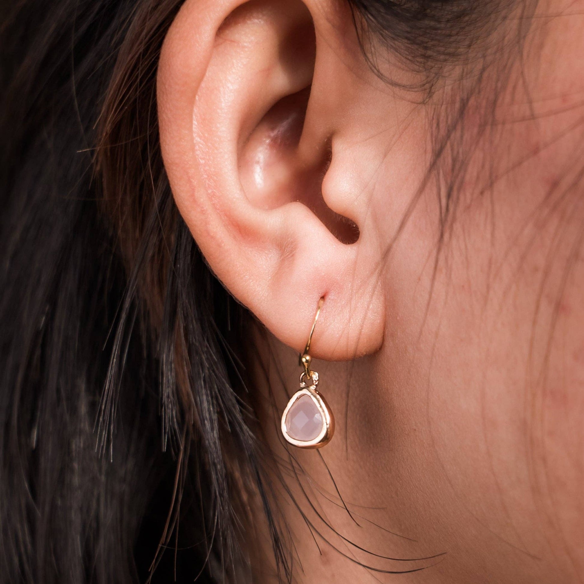 A close-up of a person's ear wearing a delicate gold dangle earring with a teardrop-shaped pale pink stone- Desert Sunrise.