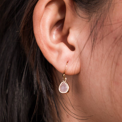 A close-up of a person's ear wearing a delicate gold dangle earring with a teardrop-shaped pale pink stone- Desert Sunrise.