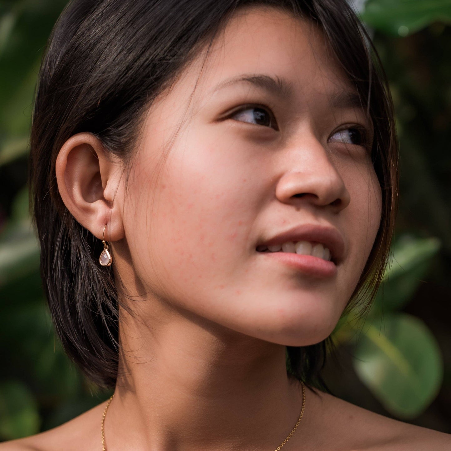 A close-up portrait of a woman with short dark hair wearing a small gold drop earring with a teardrop-shaped pink glass stone, set against a soft-focus background of green foliage.