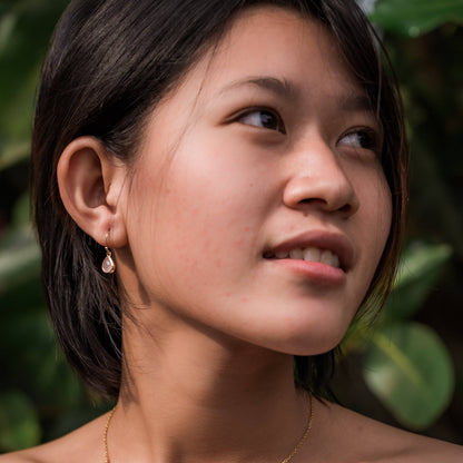 A close-up portrait of a woman with short dark hair wearing a small gold drop earring with a teardrop-shaped pink glass stone, set against a soft-focus background of green foliage.
