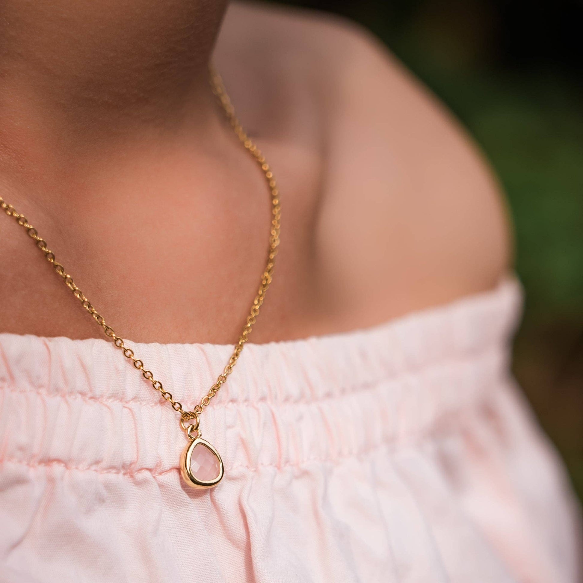 A close-up lifestyle shot of a person wearing a delicate gold chain necklace with a small pink glass stone teardrop-shaped pendant, paired with a light pink off-the-shoulder top.