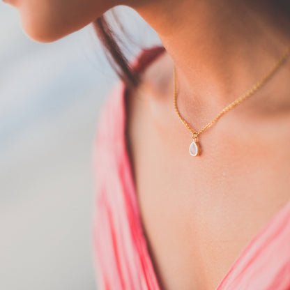 A close-up lifestyle shot of a woman wearing a delicate gold chain necklace with a small teardrop-shaped pink pendant, paired with a pink top.