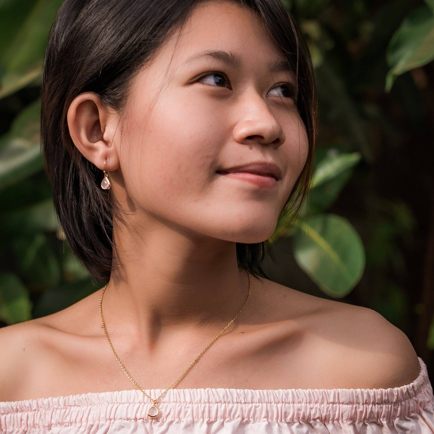 A woman with short dark hair wearing a Desert Sunrise delicate gold necklace and earring, photographed outdoors against a blurred background of green foliage.
