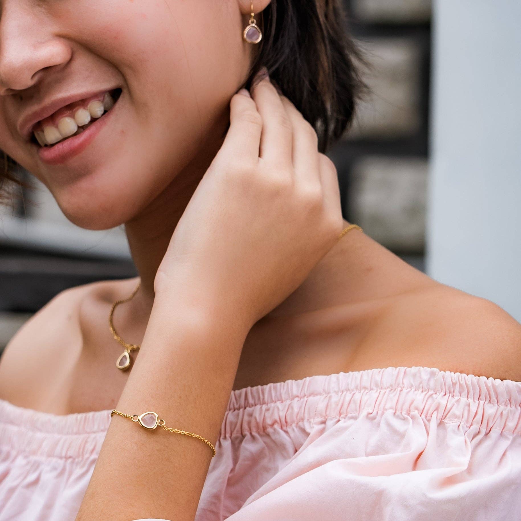 A close-up lifestyle shot of a woman wearing a delicate gold bracelet with a small pink stone on her wrist, along with matching rose quartz drop earrings and a necklace-Desert Sunrise collection.