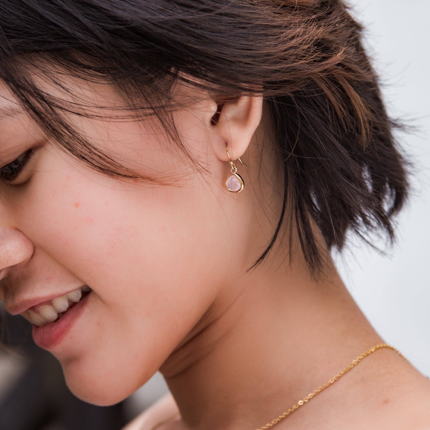 A close-up lifestyle shot of a woman wearing a delicate gold dangle earring with a round pale pink stone, paired with a fine gold chain necklace.