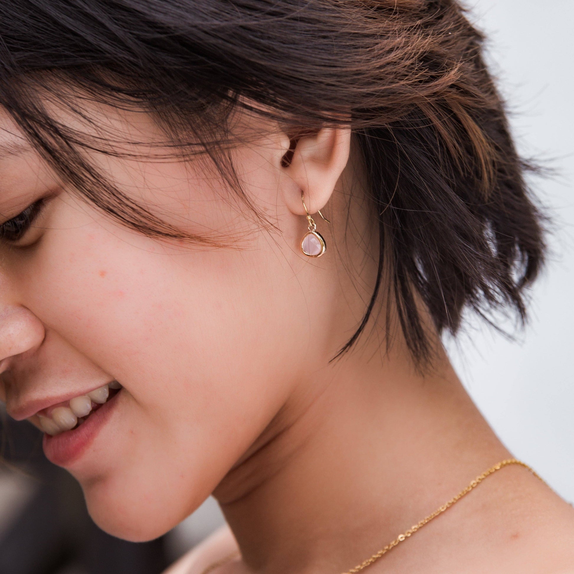 A close-up lifestyle shot of a woman wearing a delicate gold dangle earring with a round pale pink stone, paired with a fine gold chain necklace.