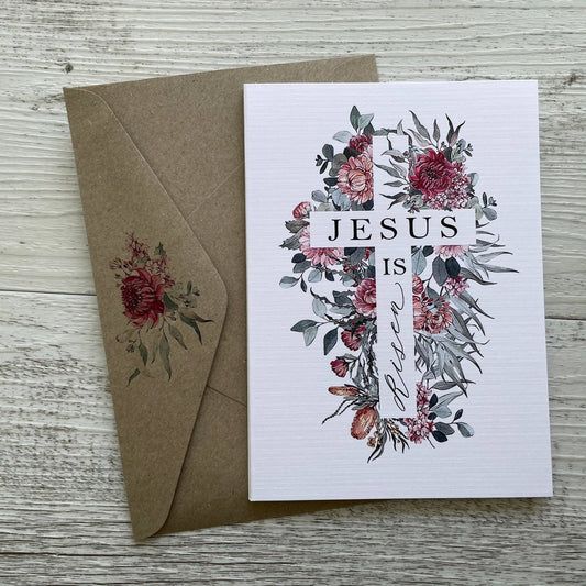 Greeting card with floral design of a cross and florals and 'Jesus is Love' text, accompanied by a brown envelope on a wooden surface.