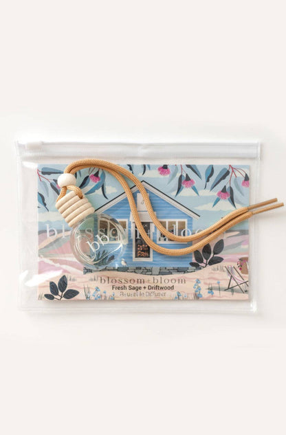 Car air freshener featuring a glass reusable diffuser bottle with a neutral tie and wooden detail displayed on a colourful background with floral patterns and the label 'Fresh Sage + Driftwood.'