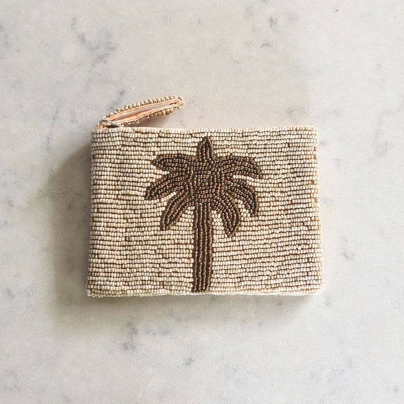 A small rectangular beaded pouch or coin purse in warm beige with a dark brown palm tree motif on the front and a metallic zipper at the top, photographed on a light marble surface.