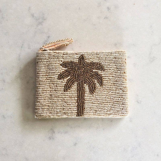 A small rectangular beaded pouch or coin purse in warm beige with a dark brown palm tree motif on the front and a metallic zipper at the top, photographed on a light marble surface.