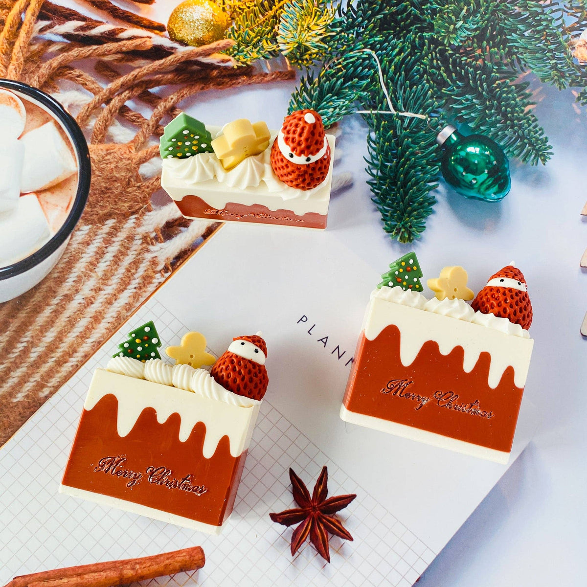 Three Handmade Artisan Christmas Soaps | Christmas Fantasia | Champagne & Strawberry Scent— arranged on a tabletop with pine branches, ornaments, a cinnamon stick, star anise, and a bowl of marshmallows. The pieces are brown with cream-colored frosting details and topped with miniature trees, stars, and Santa figures.