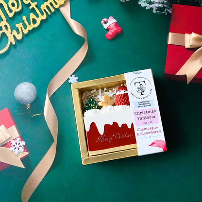 An open gold gift box containing a small Christmas-themed dessert or treat box labeled 'Christmas Fantasia' by Tammi Home Body & Bath, styled on a teal background with ribbon, ornaments, and gift boxes.
