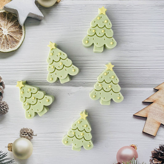 Five Handmade Artisan Christmas Soap | Jolly Evergreen | Fir Needle, Cedarwood & Patchouli scent—light green Christmas tree-shaped items with yellow star tops and dotted trim details, arranged on a light gray wooden surface with holiday props including ornaments, a dried orange slice, pinecones, and wooden cutouts.
