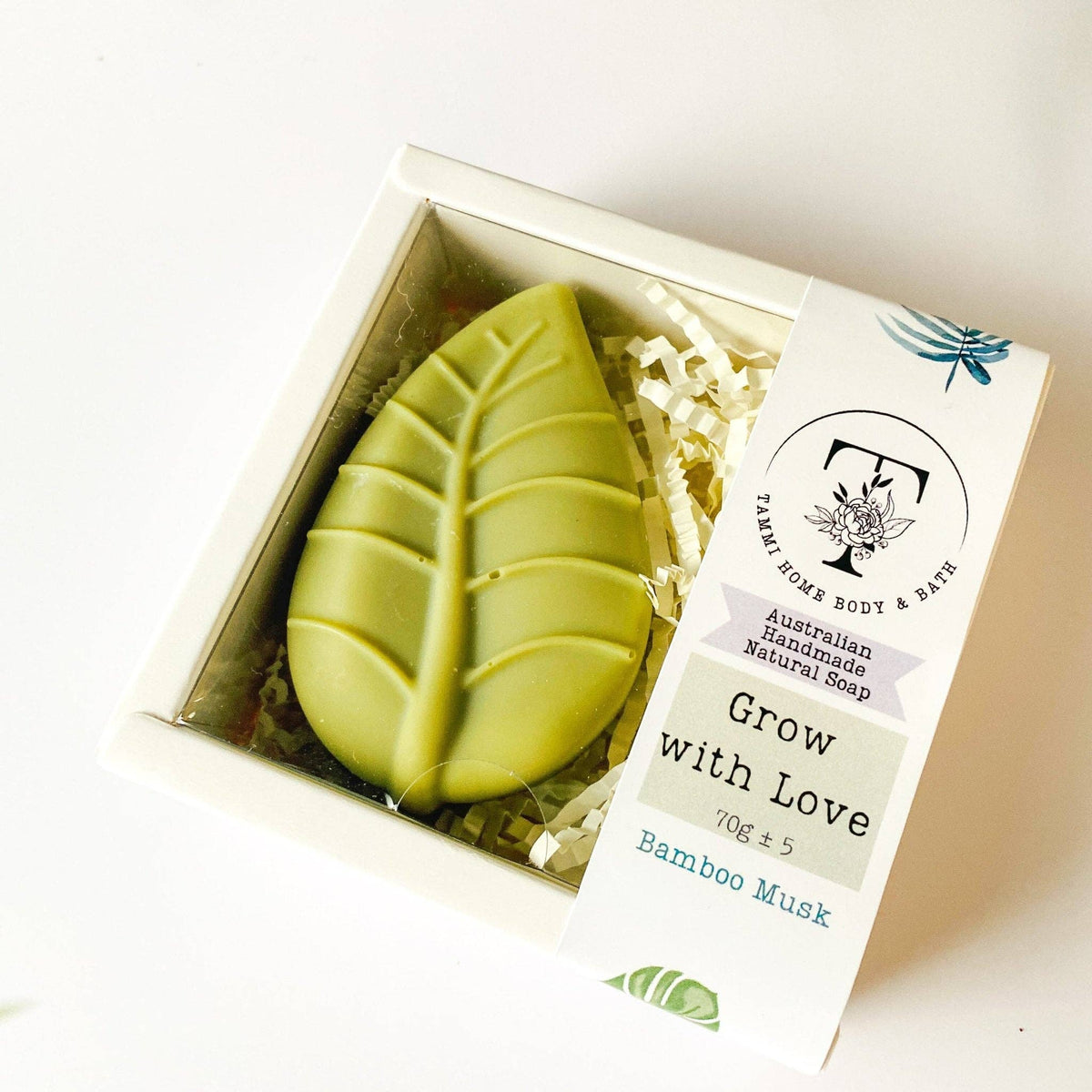 Handmade Artisan Soap | Grow with Love | Bamboo Musk Scent. A leaf-shaped green soap bar in a clear-lidded white gift box with shredded paper filler and a paper label reading 'Grow with Love' and 'Bamboo Musk' from Palm Home Body & Bath.
