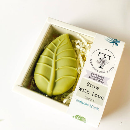 Handmade Artisan Soap | Grow with Love | Bamboo Musk Scent. A leaf-shaped green soap bar in a clear-lidded white gift box with shredded paper filler and a paper label reading 'Grow with Love' and 'Bamboo Musk' from Palm Home Body & Bath.
