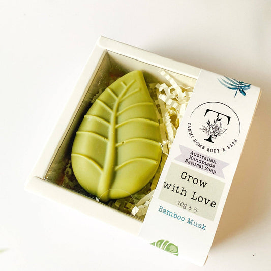 Handmade Artisan Soap | Grow with Love | Bamboo Musk Scent. A leaf-shaped green soap bar in a clear-lidded white gift box with shredded paper filler and a paper label reading 'Grow with Love' and 'Bamboo Musk' from Palm Home Body & Bath.