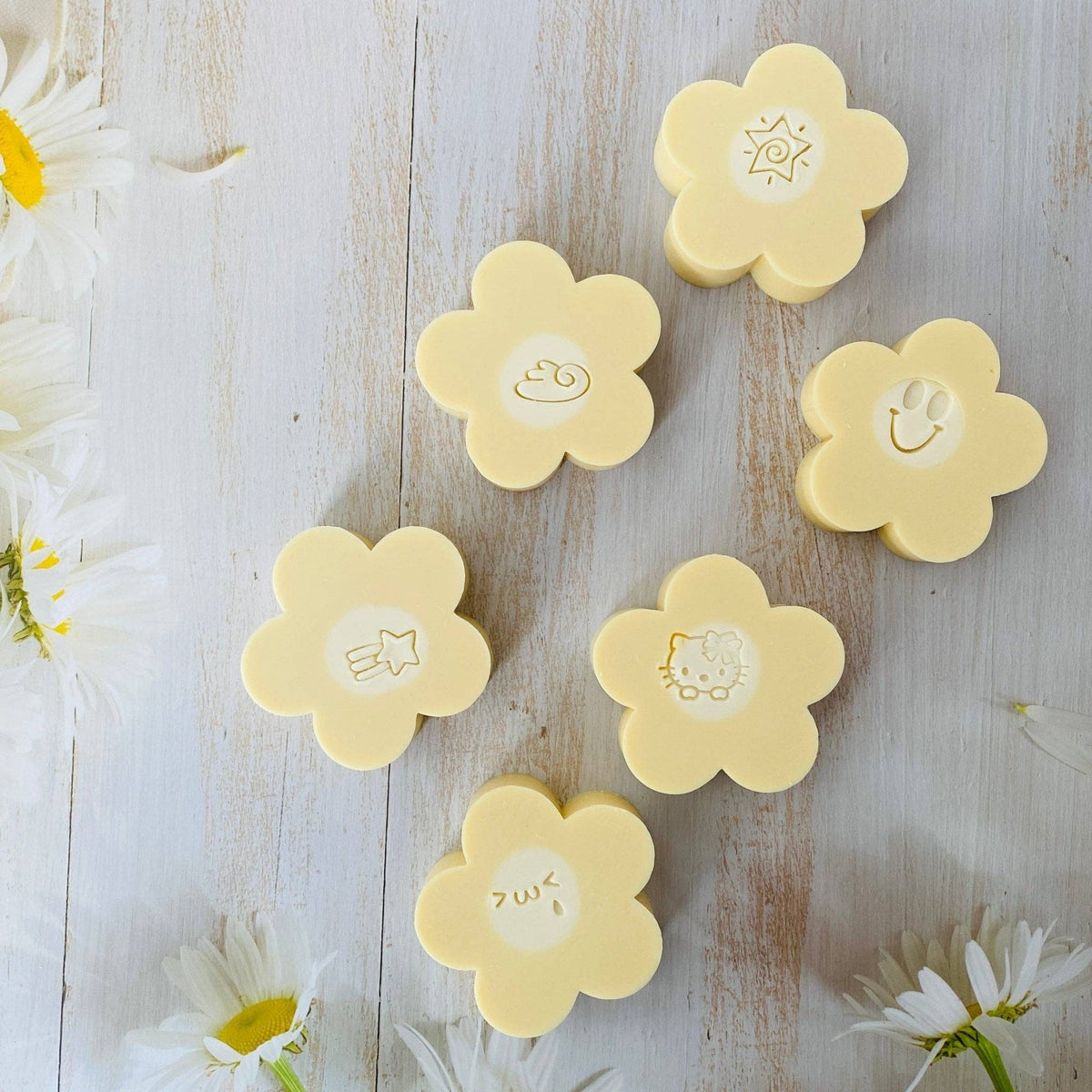 Handmade Artisan Soap with Silk | Joyful Petals | Australian Bush Flowers Scent—Six pale yellow flower-shaped soap bars arranged on a whitewashed wooden surface with white daisies around the edges. Each soap has a different simple embossed face or symbol in the center.