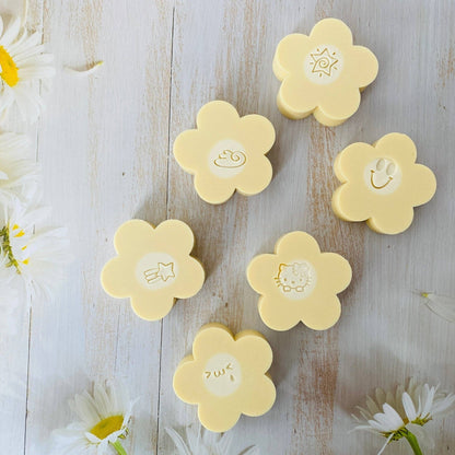 Handmade Artisan Soap with Silk | Joyful Petals | Australian Bush Flowers Scent—Six pale yellow flower-shaped soap bars arranged on a whitewashed wooden surface with white daisies around the edges. Each soap has a different simple embossed face or symbol in the center.