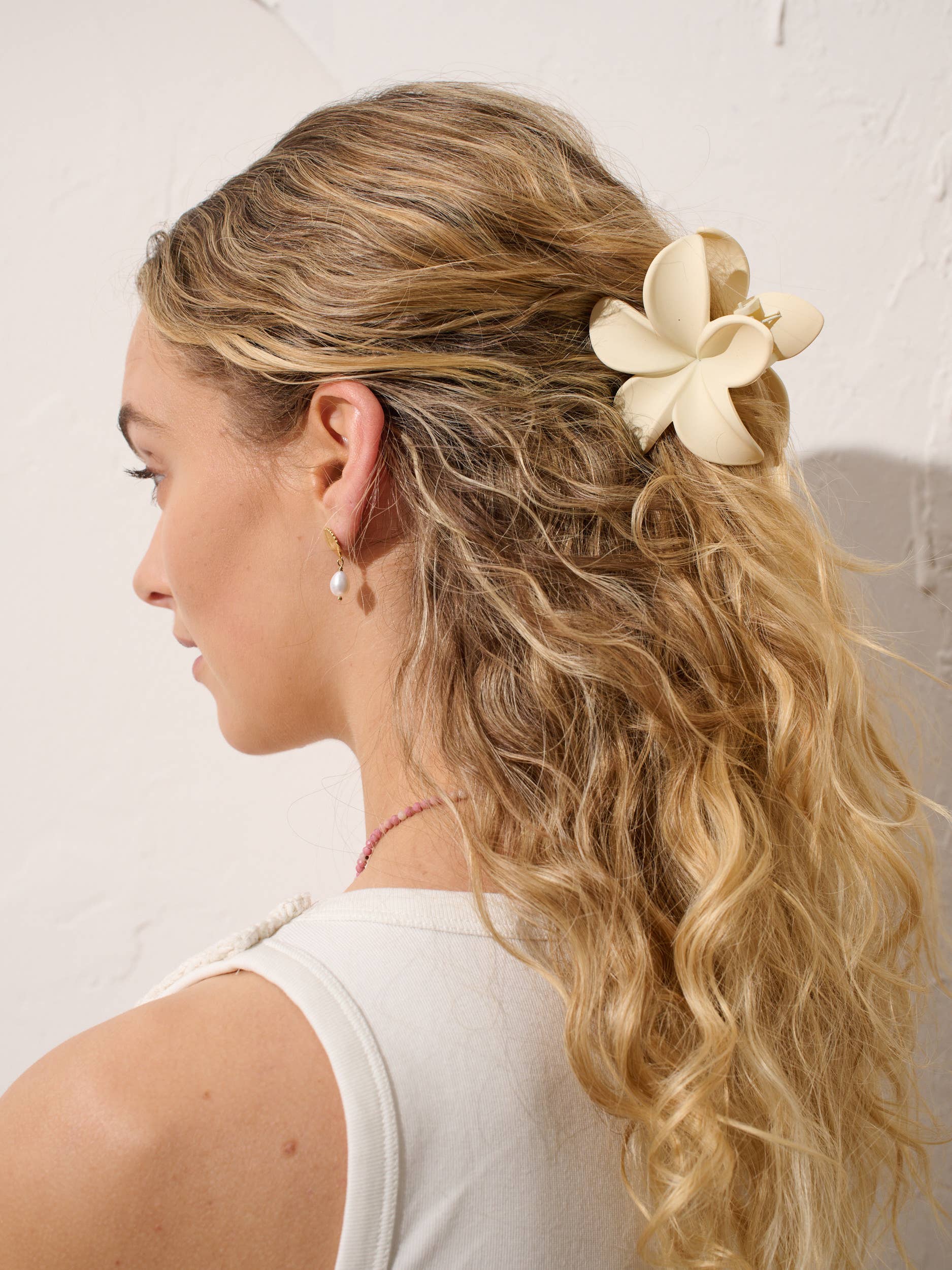 Woman with half up blonde hairstyle with hibiscus white hair claw clip, pearl and gold earrings and beaded pink necklace against a white textured backdrop.
