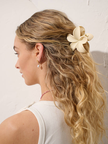 Woman with half up blonde hairstyle with hibiscus white hair claw clip, pearl and gold earrings and beaded pink necklace against a white textured backdrop.