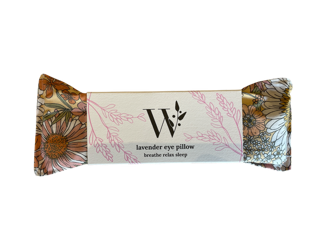 A lavender eye pillow with a daisy floral print in orange, yellow, white and pink tones. The pillow has a white label in the center with a decorative pink botanical border, displaying a stylized "W" logo for Wheelers Hills Soaps. Also has the text "lavender eye pillow-breather relax sleep."