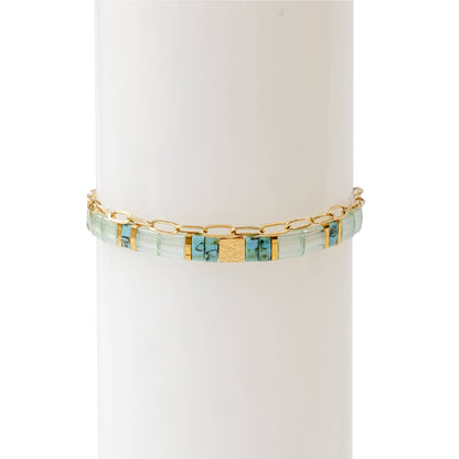 A two-piece bracelet stack displayed on a white cylindrical stand. The first bracelet features hand-strung rectangular glass beads in soft turquoise, blue, and gold tones, with central 18K gold-plated stainless steel bead engraved with a delicate leaf motif. The second bracelet is a thin, 18K gold-plated stainless steel oval chain, adding an elegant and minimal touch The combination of colours and textures creates a refined yet artistic look.
