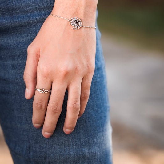 A close-up of a person's hand wearing a delicate silver chain bracelet with a circular geometric sunburst charm and a matching Olive Branch silver ring on the finger, paired with blue denim clothing against a blurred outdoor background.