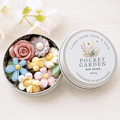 A round metal tin of Tammi Home Body & Bath Pocket Garden Mini Soaps, shown open with colorful flower-shaped soaps inside next to the lid, on a light wood surface with soft white flowers blurred in the background.