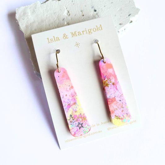 Handmade pink and purple resin dangle earrings with sparkles, on a display card from Isla & Marigold.