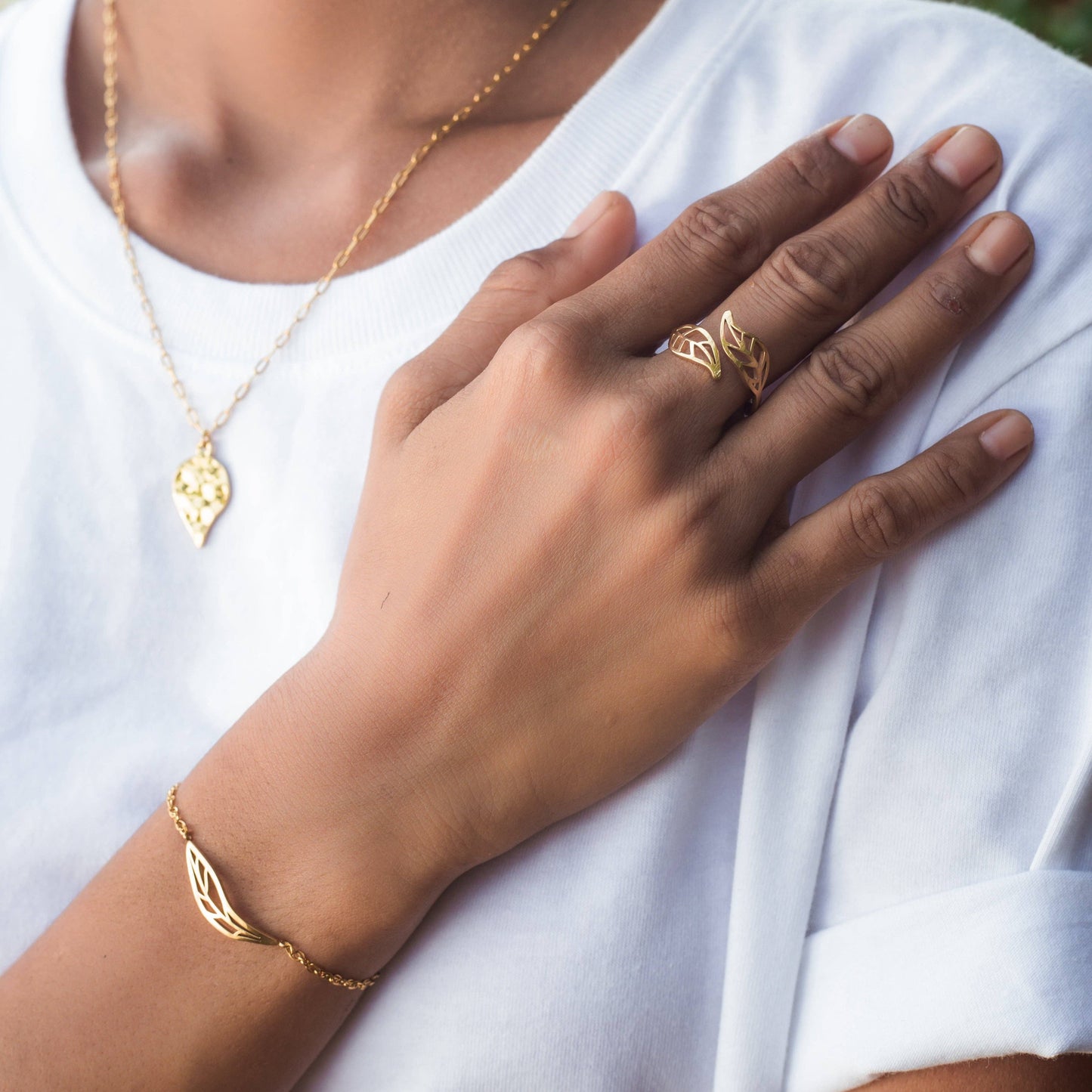 Close-up of a hand wearing a gold ring, bracelet and necklace with a leaf design on a white background.