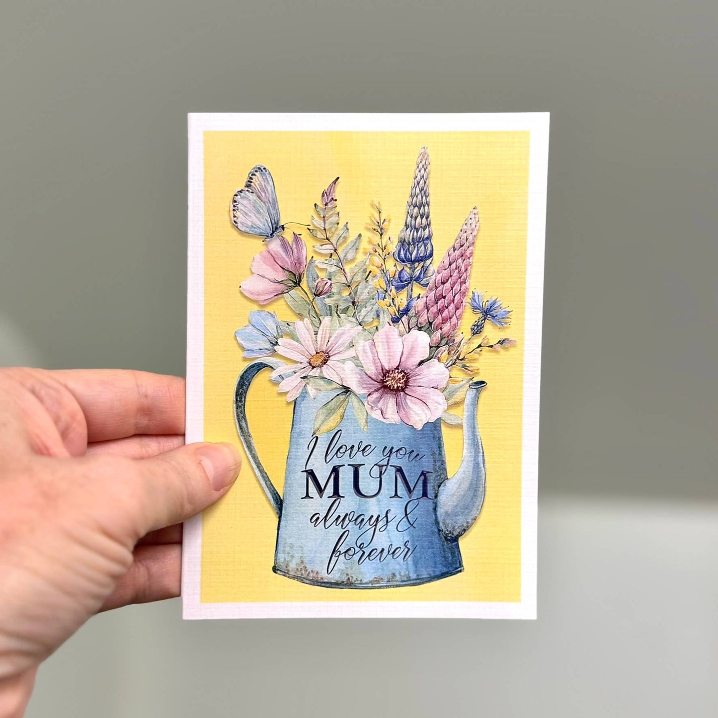 Greeting card with floral design and 'I love you Mum always & forever' text, held by a hand against a neutral background.