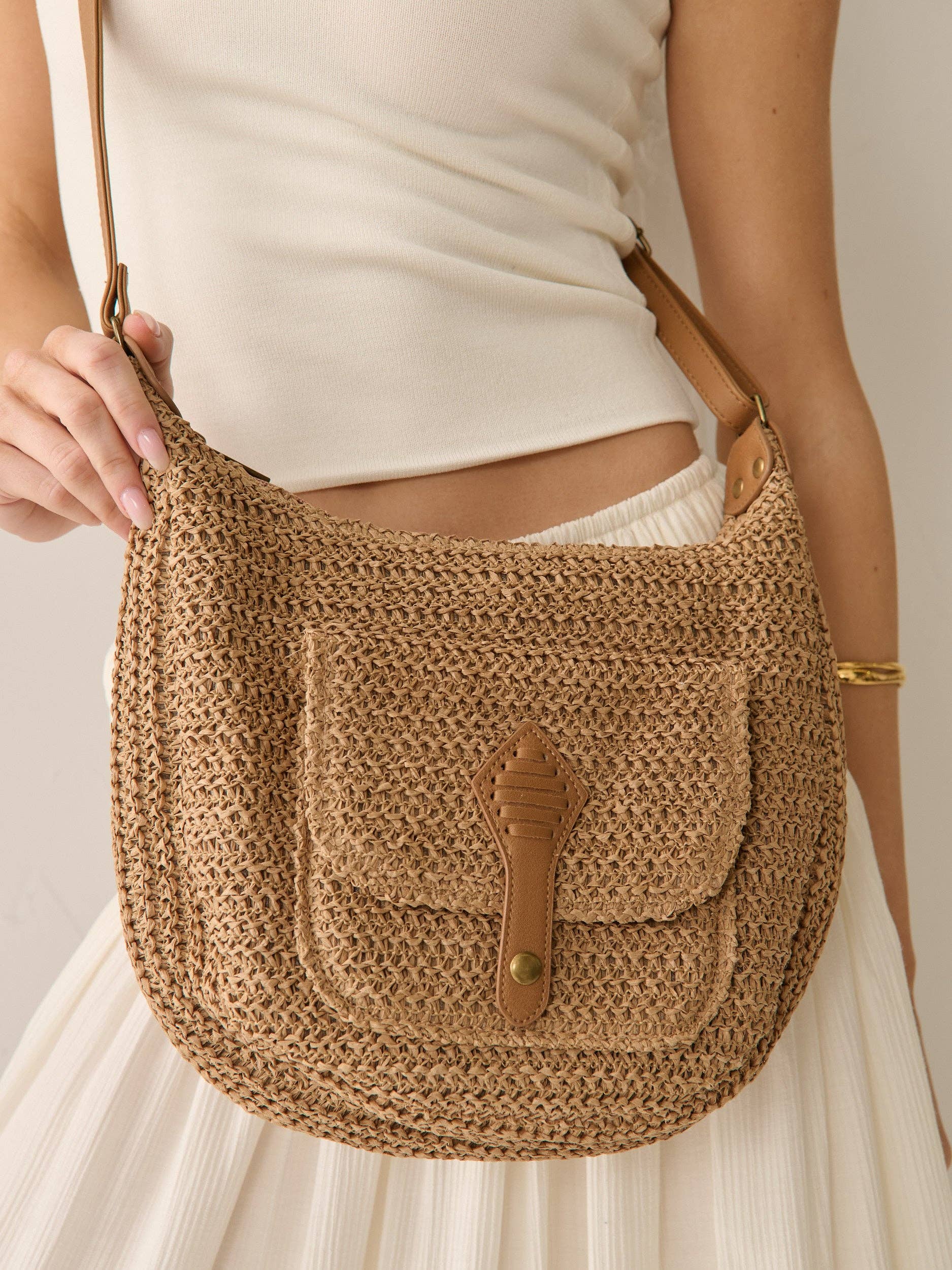 Brown crochet style texture crossbody handbag held by a person wearing a white top and skirt against a neutral background