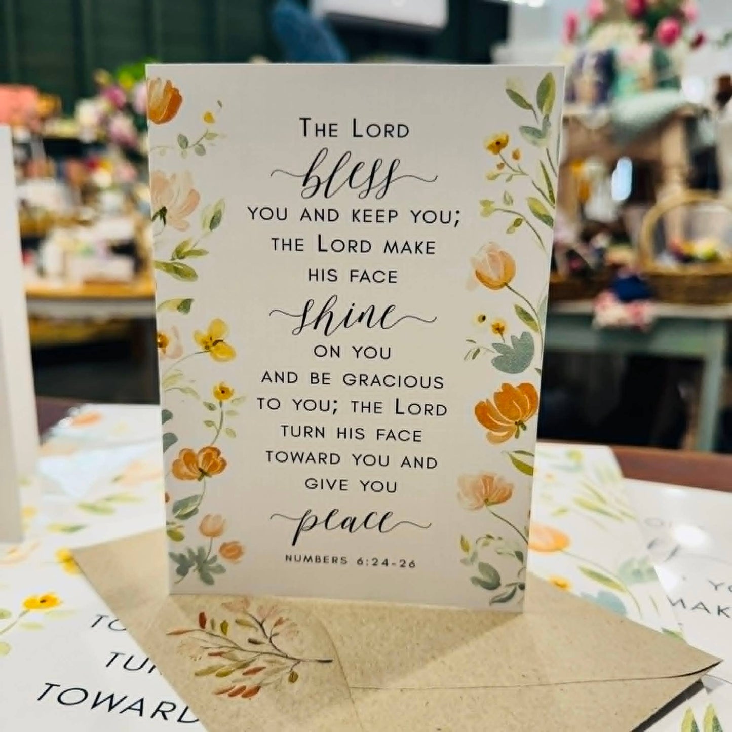 A vertical printed card with floral illustrations and a Bible verse, standing upright on a table in a blurred shop setting. The card reads: 'The Lord bless you and keep you; the Lord make his face shine on you and be gracious to you and give you place.
