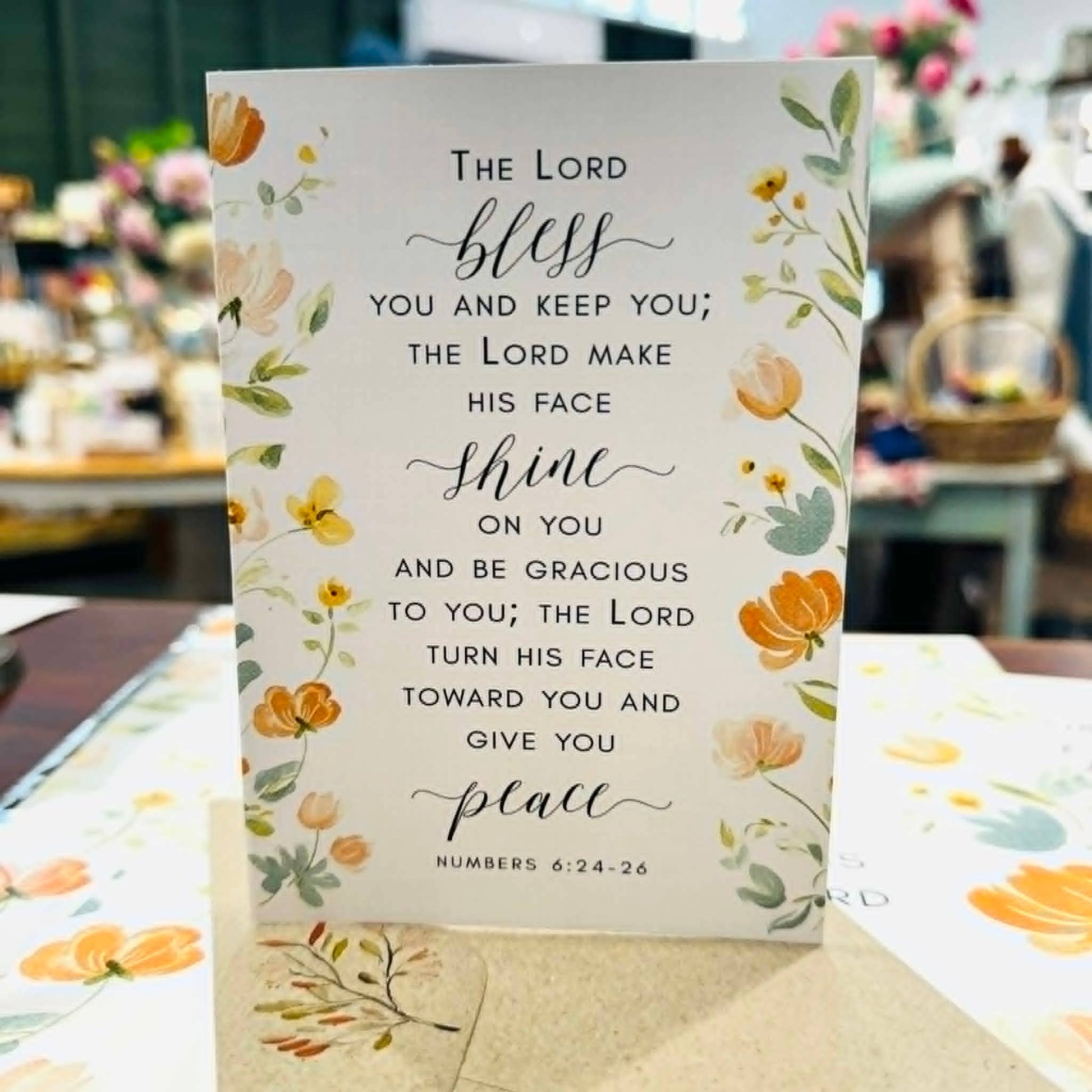 A vertical white prayer card with floral illustrations in yellow, orange, and green, featuring a Bible verse from Numbers 6:24-26. The card is standing upright on a table with a matching floral envelope in front of a softly blurred shop background.