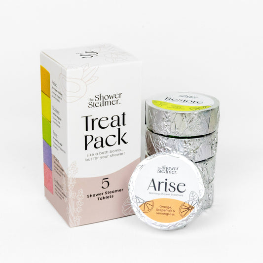 The Shower Steamer Treat Pack featuring a white and pink outer box, a stack of silver foil-wrapped round containers, and one round container labeled 'Arise' in the foreground, all on a clean white background.