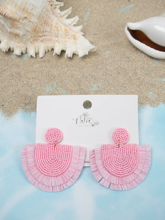 Soft baby pink beaded raffia earrings on a Nakoa jewellery card. Sand and shells in the background.
