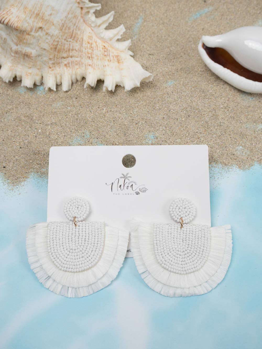 A pair of white beaded raffia earrings on a Nakoa branded jewellery card. Sand and shells in the background.