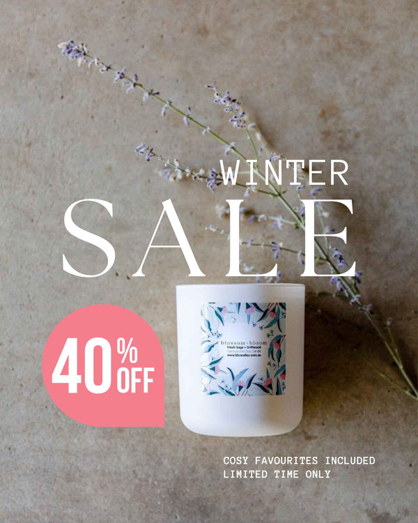 A Blossom + Bloom white scented candle jar with a botanical label sits on a textured beige surface with dried lavender sprigs. Large overlaid text reads 'WINTER SALE' with a pink circle saying '40% OFF', and smaller text at the bottom says 'COSY FAVOURITES INCLUDED LIMITED TIME ONLY'.