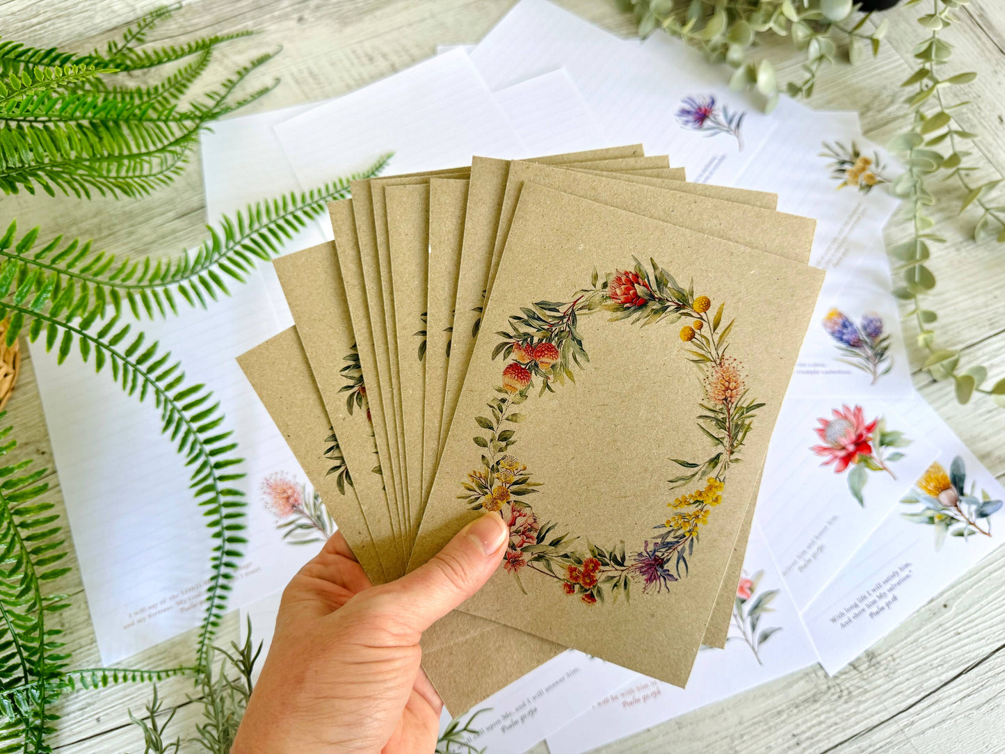 Hand holding a stack of brown paper kraft envelopes with floral wreath designs on a white surface with greenery.